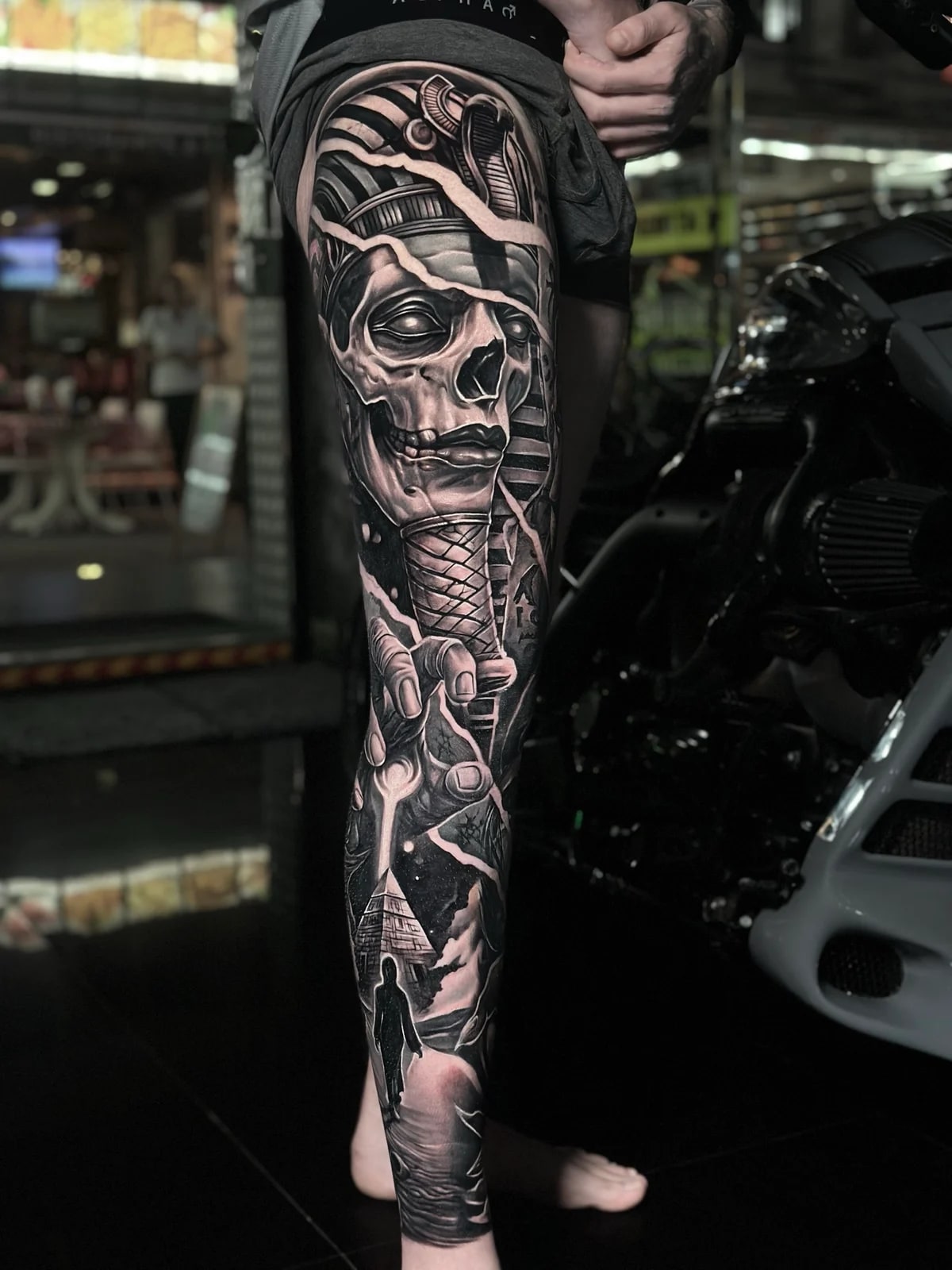Detailed black and grey realism full leg sleeve tattoo featuring a fragmenting Egyptian pharaoh skull, reaching hand, and pyramid under a starry sky at Bloodline Tattoo Phuket.