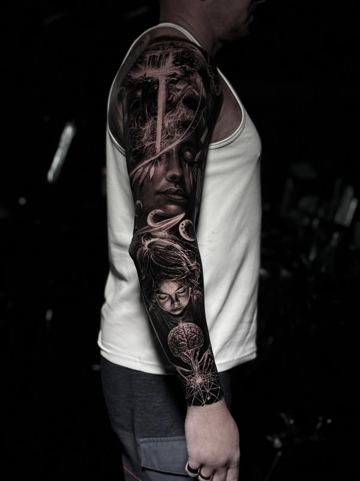 Detailed black and grey realism full sleeve tattoo featuring a serene woman with radiant cross, Saturn, child portrait, and brain in geometric pattern at Bloodline Tattoo Phuket.