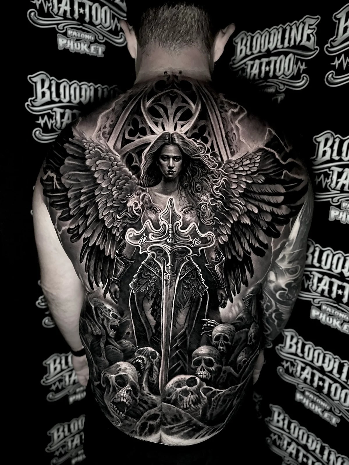 Detailed black and grey realism full back tattoo featuring a winged angel warrior with broadsword, gothic window, and pile of skulls at Bloodline Tattoo Phuket.