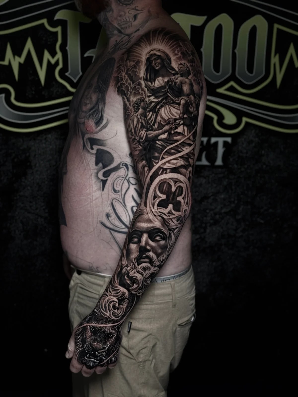 Detailed black and grey realism full sleeve tattoo featuring the Virgin Mary with infant Jesus, gothic rose window, bearded statue face, and lion on the hand at Bloodline Tattoo Phuket.