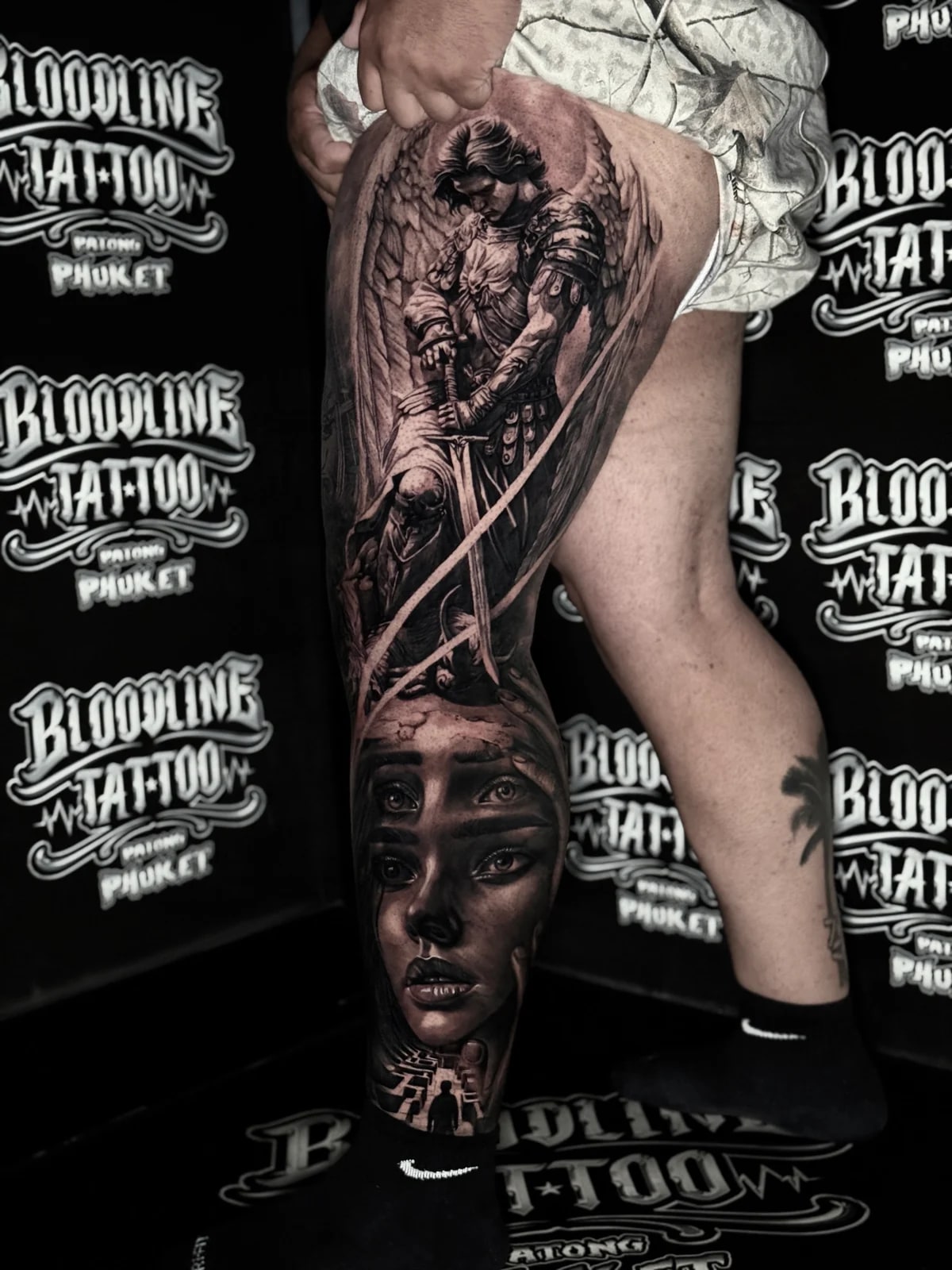 Detailed black and grey realism full leg sleeve tattoo featuring an armored angel with sword, four-eyed woman portrait, and maze silhouette at Bloodline Tattoo Phuket.