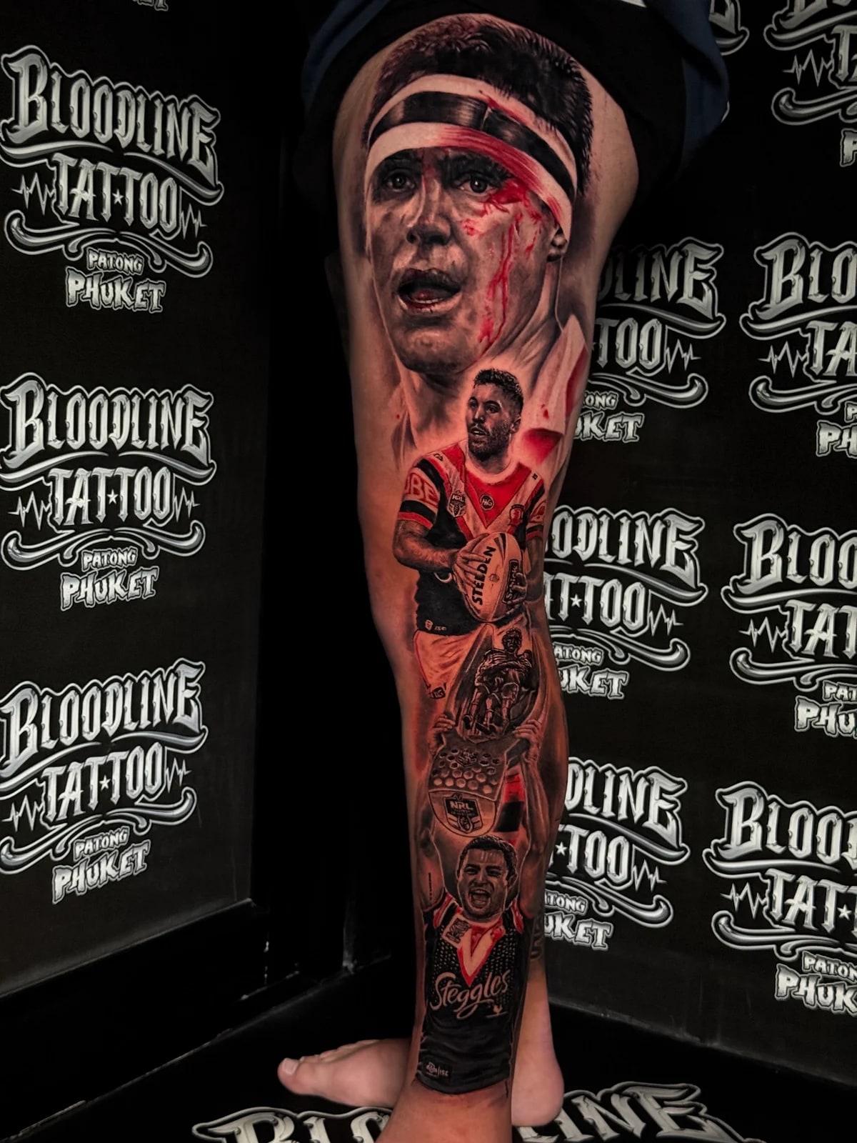 Detailed black and grey realism full leg sleeve tattoo with red accents featuring Sydney Roosters rugby players, Steeden ball, and NRL trophy at Bloodline Tattoo Phuket.