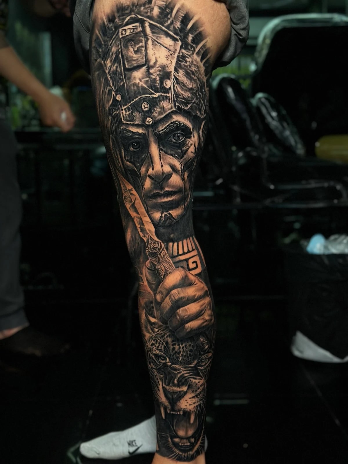 Detailed black and grey realism full leg sleeve tattoo featuring an indigenous warrior in an ornate headdress, hand gripping a decorative dagger, and snarling jaguar at Bloodline Tattoo Phuket.