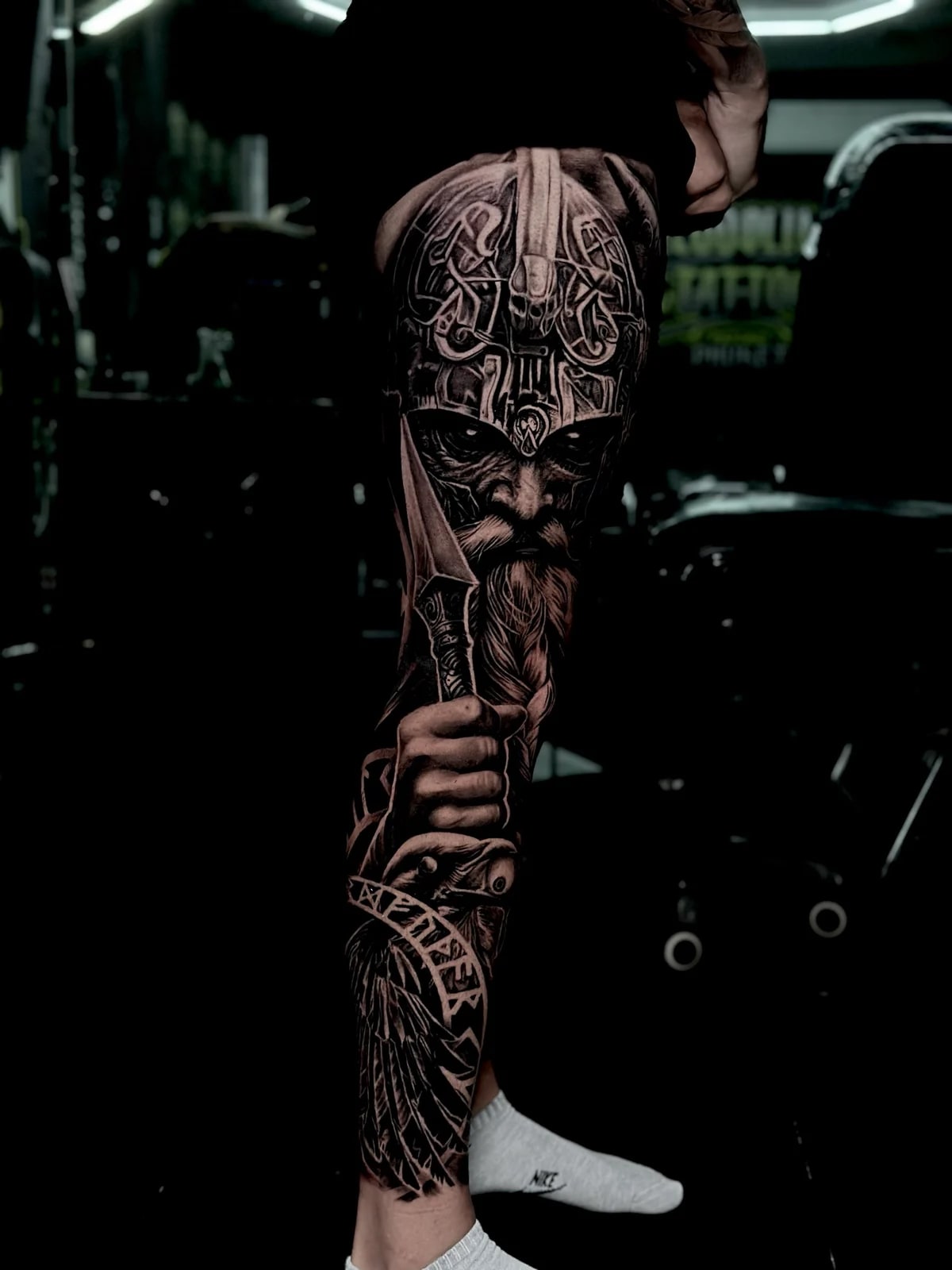 Intricate black and grey realism full leg sleeve tattoo featuring a Viking knotwork helmet, braided warrior beard, hand on sword, raven, and runic banner at Bloodline Tattoo Phuket.