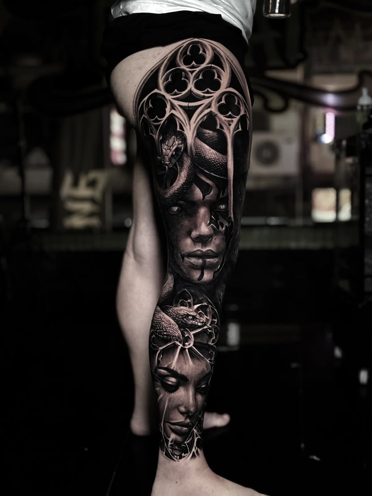 Detailed black and grey realism full leg sleeve tattoo featuring gothic cathedral arches, two haunting women's portraits, and coiled snakes at Bloodline Tattoo Phuket.