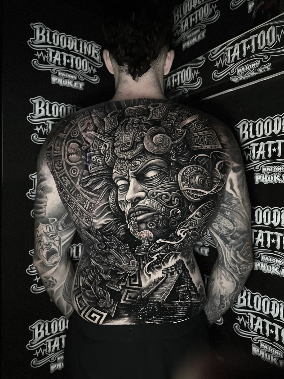 Intricate black and grey realism full back tattoo featuring a Mayan warrior with ornate feathered headdress, stepped pyramid, and feathered serpent skull at Bloodline Tattoo Phuket.