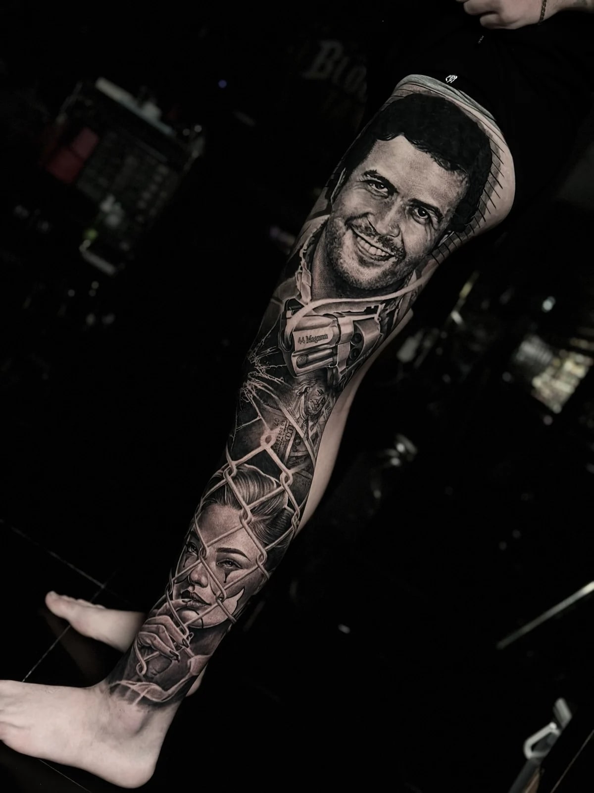 Detailed black and grey realism full leg sleeve tattoo featuring Pablo Escobar portrait, .44 Magnum revolver, hundred-dollar bills, and woman behind chain-link fence at Bloodline Tattoo Phuket.