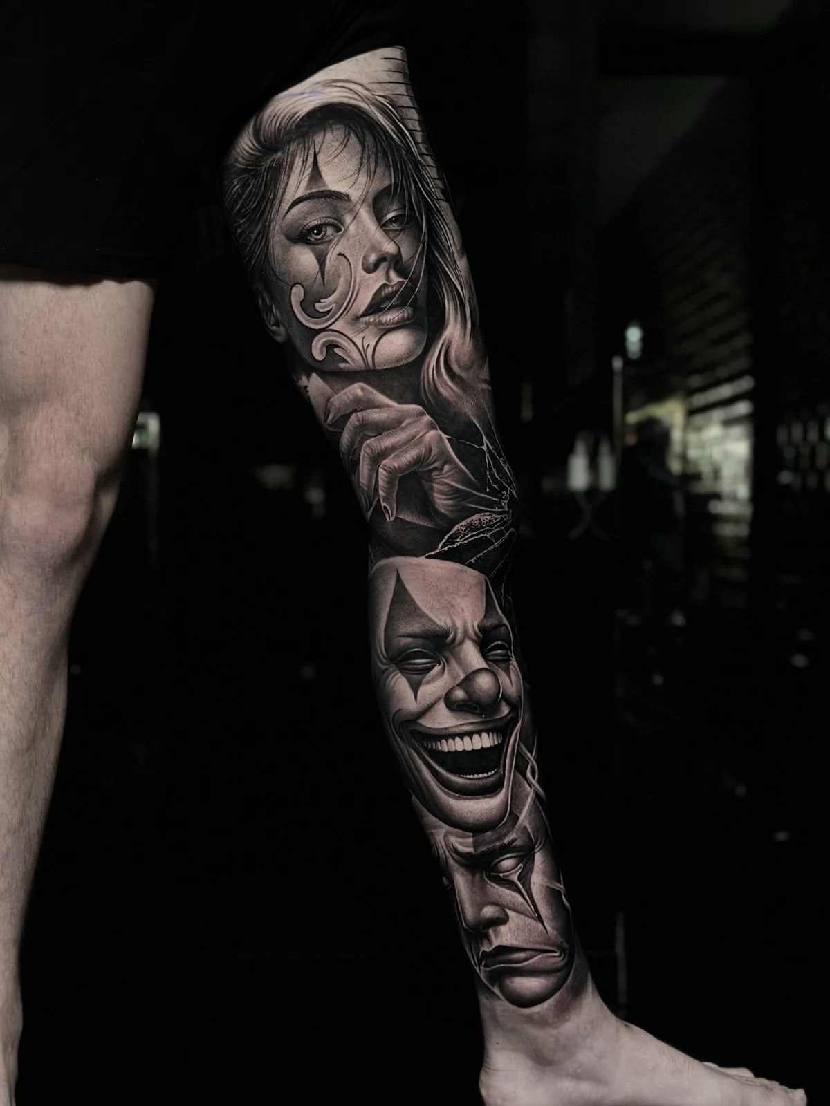 Detailed black and grey realism full leg sleeve tattoo featuring a woman with clown makeup, laughing clown mask, and sad clown mask at Bloodline Tattoo Phuket.