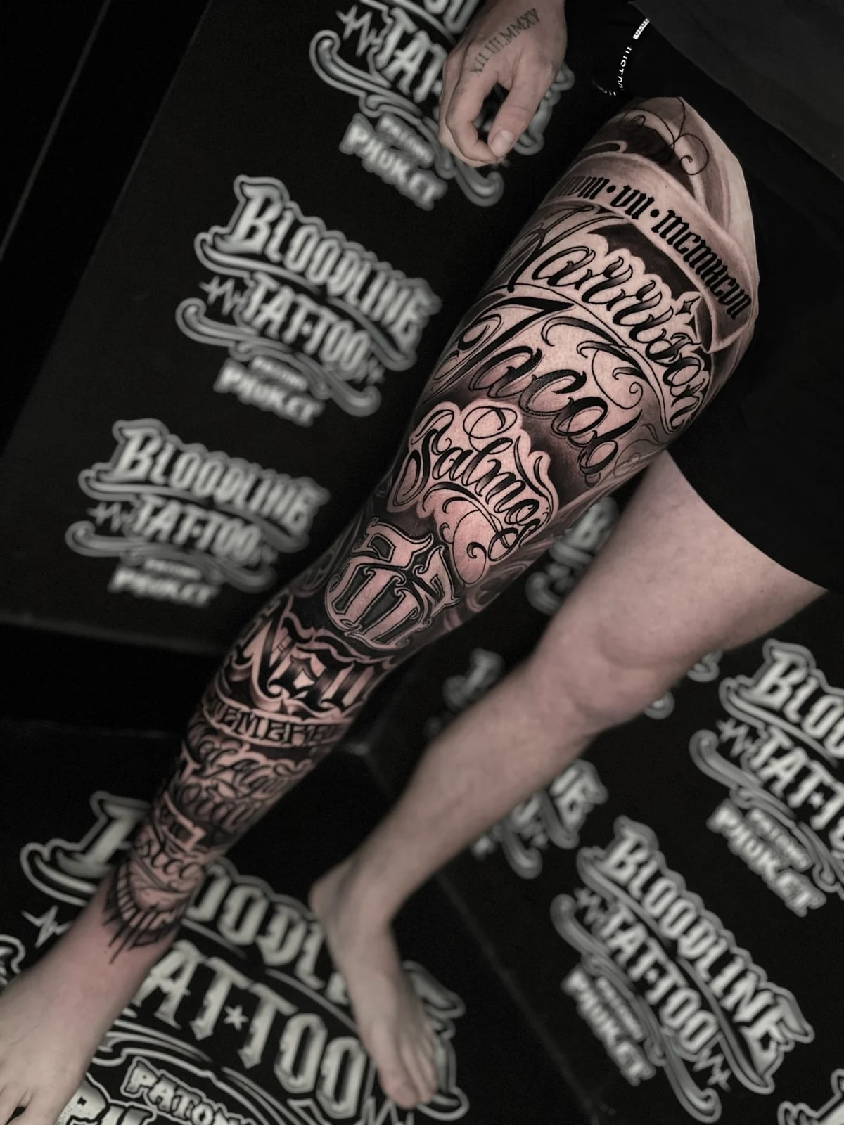 Detailed black and grey realism full leg sleeve tattoo featuring names Harrison, Jacob, Salmon, and Neil in intricate script with Roman numerals at Bloodline Tattoo Phuket.