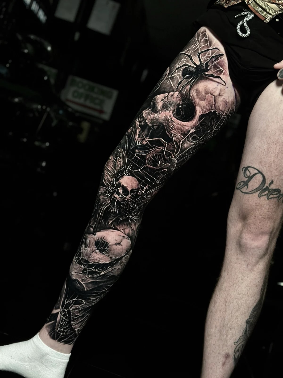Detailed black and grey realism full leg sleeve tattoo featuring multiple realistic skulls, large spider, and intricate spider webs at Bloodline Tattoo Phuket.