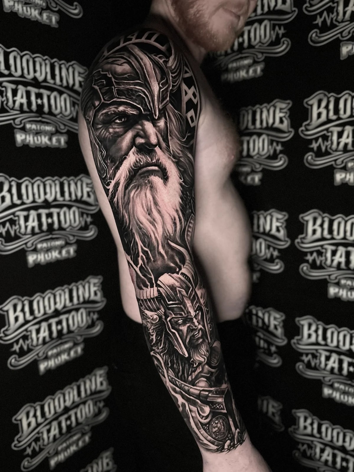 Detailed black and grey realism full sleeve tattoo featuring two Viking warriors with ornate winged helmets, beards, and lightning effects at Bloodline Tattoo Phuket.