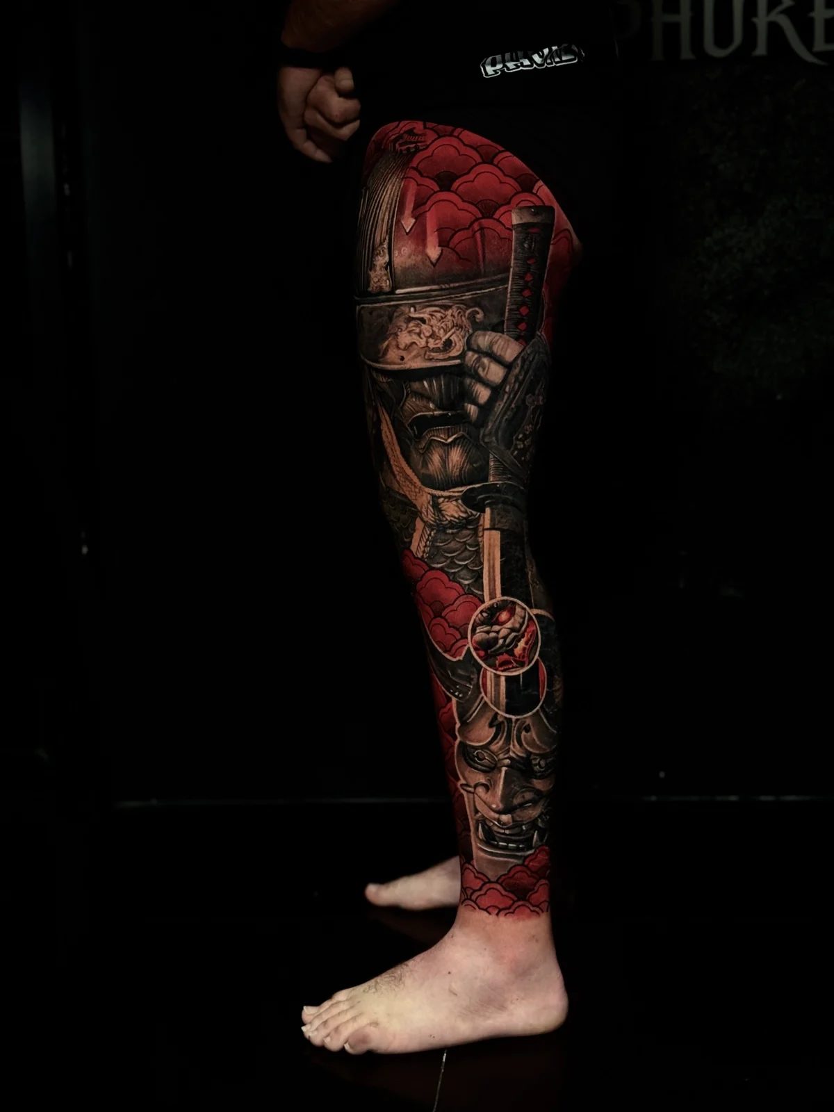 Japanese-inspired colour leg tattoo of an armored samurai with a katana and a Hannya mask amid red cloud accents at Bloodline Tattoo Phuket.