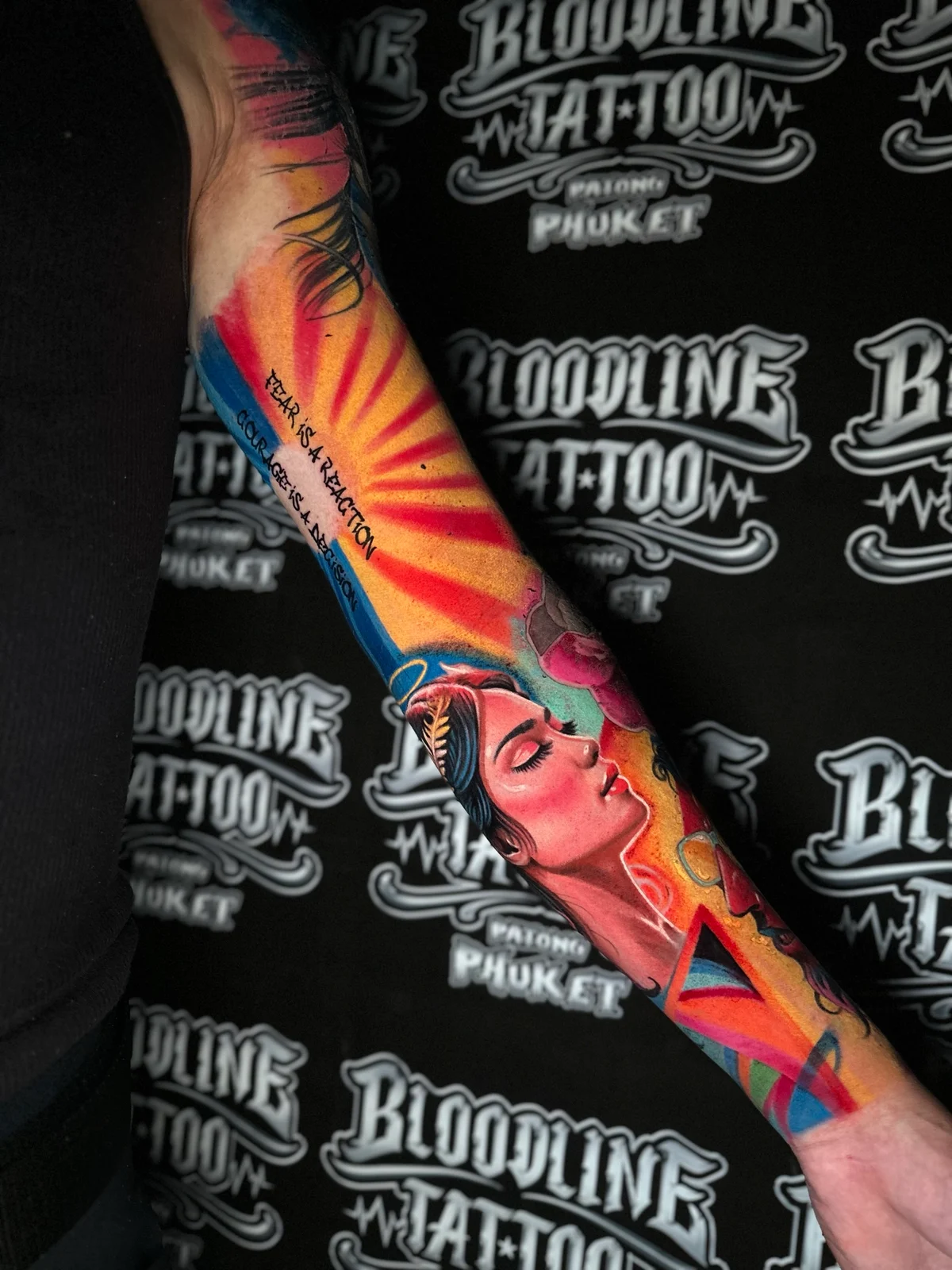 Colour forearm tattoo of a laurel-crowned woman profile with FEAR IS A REACTION and COURAGE IS A DECISION script beside a sunburst at Bloodline Tattoo Phuket.