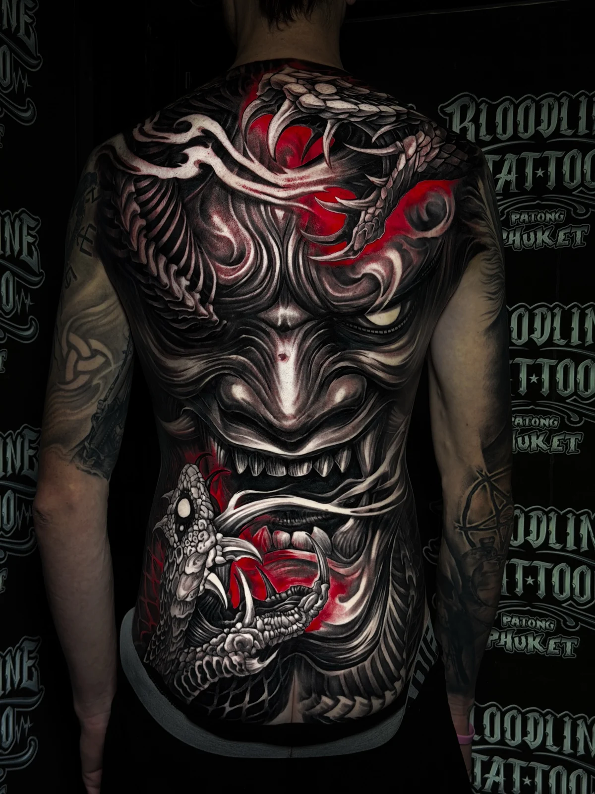 Japanese full-back Hannya mask flanked by snakes with fangs and deep red accents in black and grey at Bloodline Tattoo Phuket.