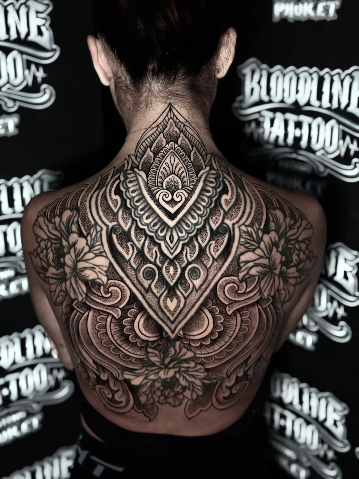 Symmetrical ornamental back mandala with a diamond central motif, stippled dotwork shading, and large peony florals on the shoulder blades at Bloodline Tattoo Phuket.