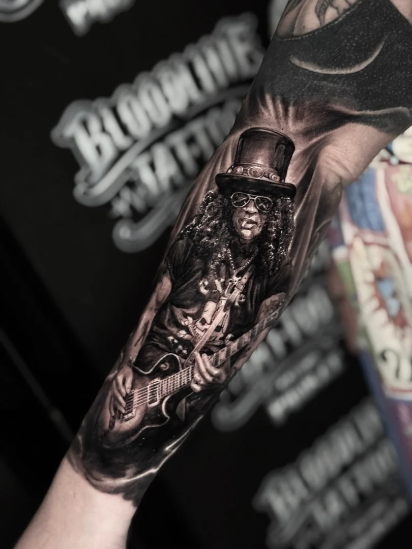 Black and grey portrait realism forearm tattoo of Slash in top hat playing a Les Paul guitar at Bloodline Tattoo Phuket.