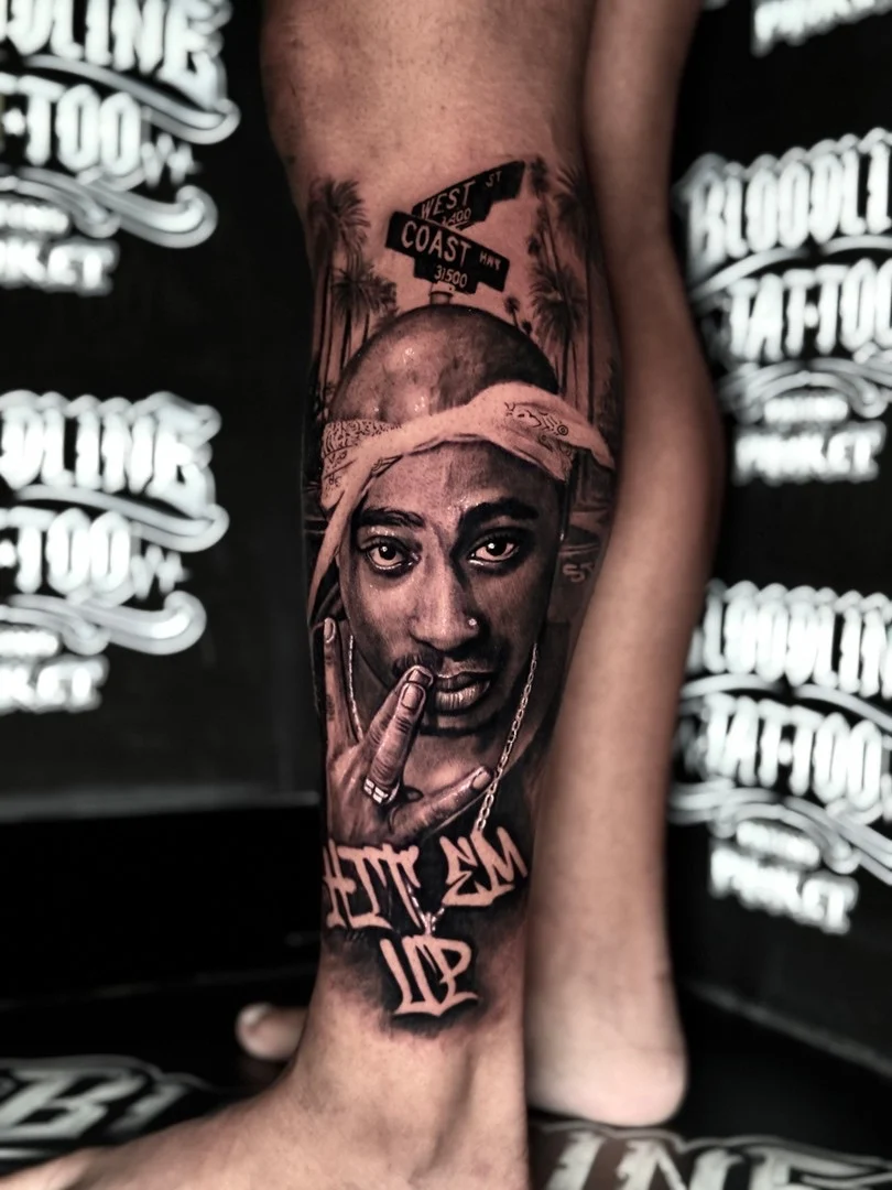 Black and grey portrait realism leg tattoo of Tupac with West St street signs and Hit Em Up script at Bloodline Tattoo Phuket.