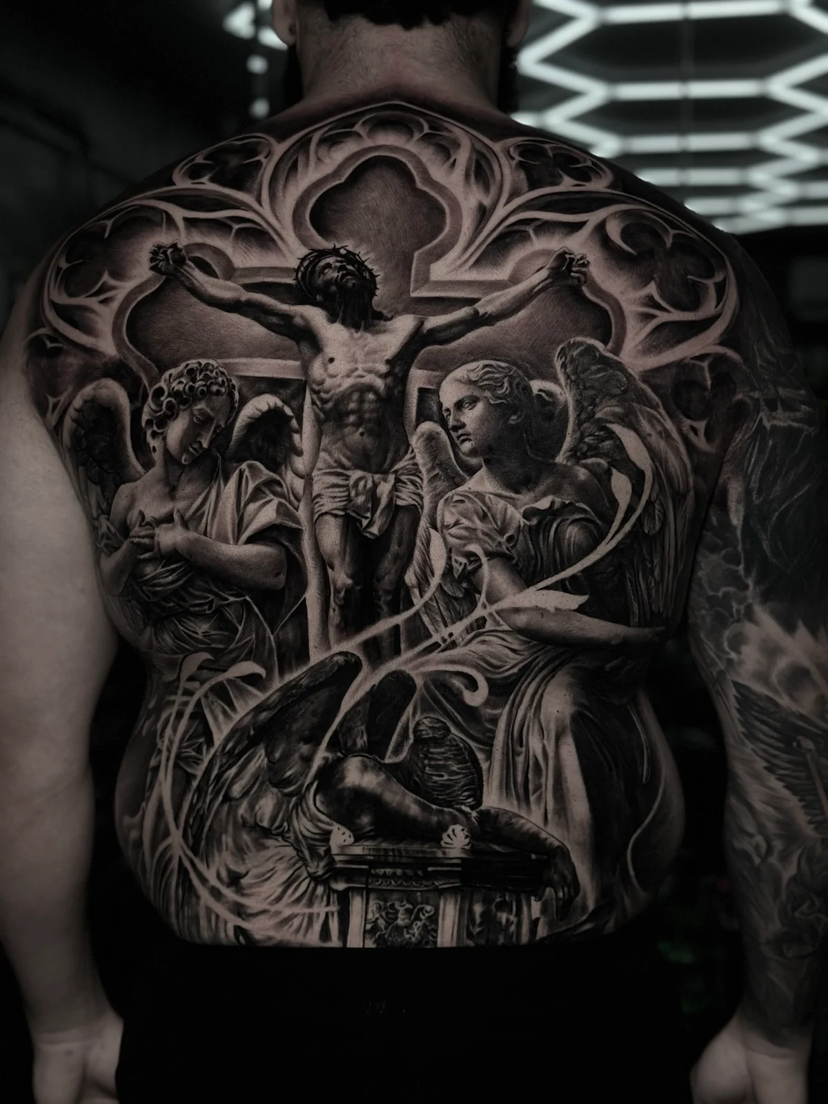Large detailed black and grey realism full back tattoo depicting the crucifixion of Jesus Christ with mourning angels and Gothic architectural framing at Bloodline Tattoo Phuket.