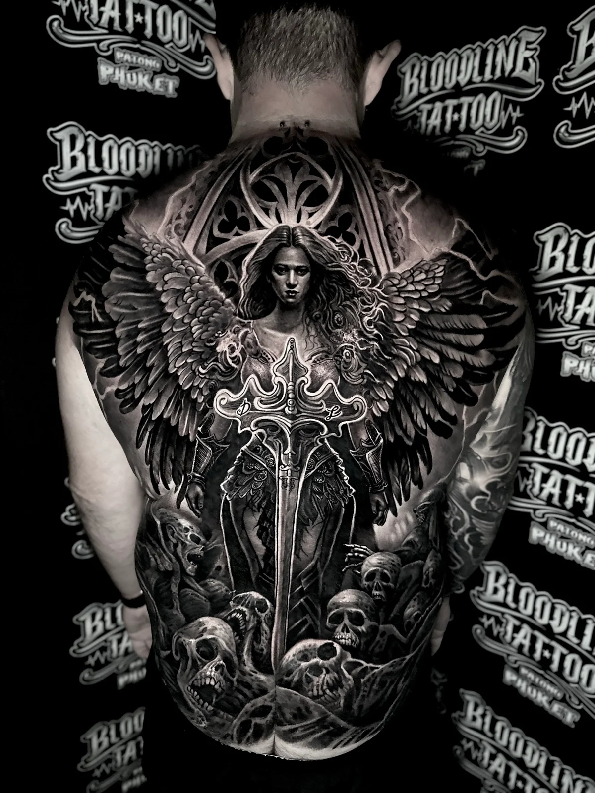 Detailed black and grey realism full back tattoo featuring a winged angel warrior with broadsword, gothic window, and pile of skulls at Bloodline Tattoo Phuket.