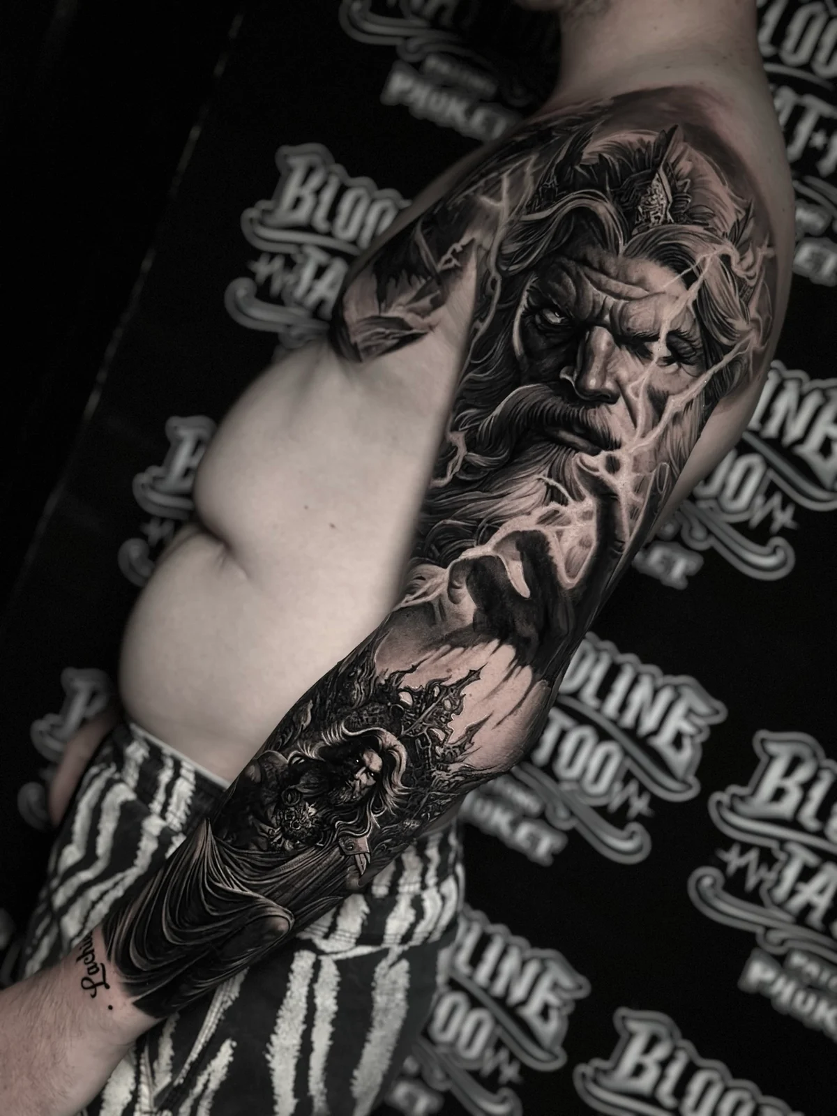 Detailed black and grey realism full sleeve tattoo featuring Zeus with lightning bolts and Greek mythology elements at Bloodline Tattoo Phuket.